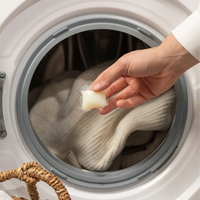 Une main place une capsule de lessive hydrosoluble dans le tambour d'un lave-linge rempli de linge.