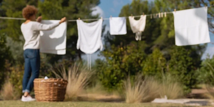 Une personne étend du linge blanc propre sur une corde dans un jardin ensoleillé à côté d'un panier en osier.
