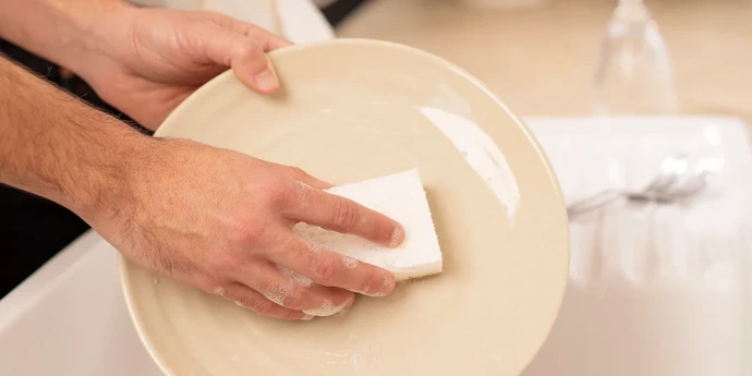 Deux mains lavent une assiette beige avec une éponge blanche moussante au-dessus d'un évier.