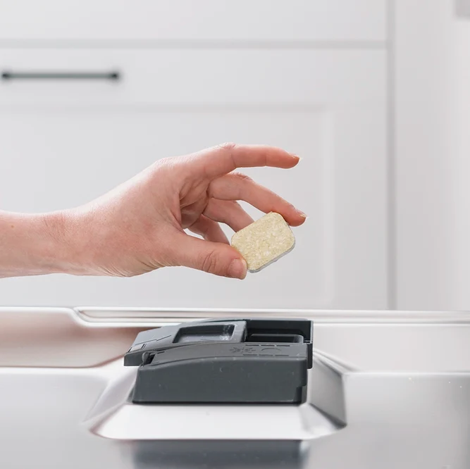 Une main place une tablette de lavage sans film plastique au-dessus du bac à produit d'un lave-vaisselle.