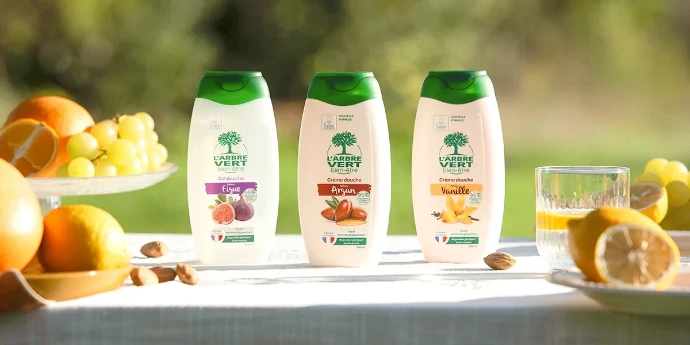Gamme Douche L'Arbre Vert bien-être : des formules hypoallergéniques au pH neutre pour nettoyer et prendre soin de la peau au quotidien. Trois flacons de gels et crèmes douche L'Arbre Vert bien-être aux parfums Figue, Argan et Vanille, alignés sur une table en extérieur entourée de fruits frais.