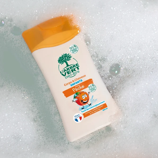 Flacon de gel douche corps et cheveux Enfants L'Arbre Vert bien-être au parfum Pêche, posé au milieu de la mousse de bain.