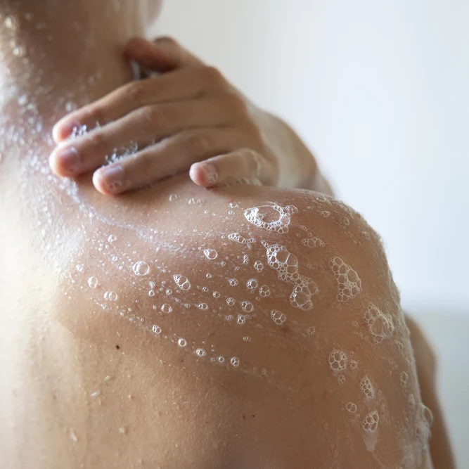 Une main appliquant de la mousse de gel douche sur une épaule et le haut du dos sous la douche.