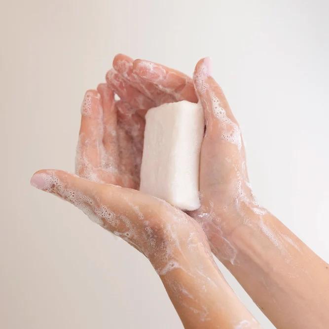 Deux mains mouillées et couvertes de mousse tenant un pain de savon solide blanc.