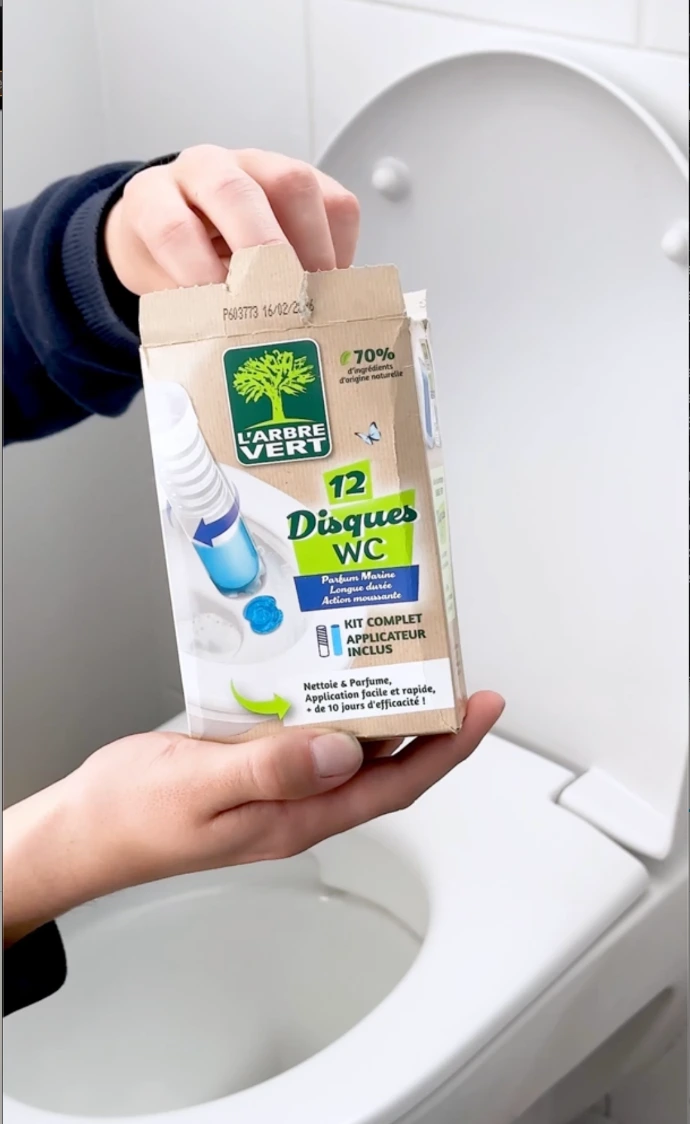Deux mains tenant un étui de 12 Disques WC L'Arbre Vert parfum Marine au-dessus d'une cuvette de toilettes.
