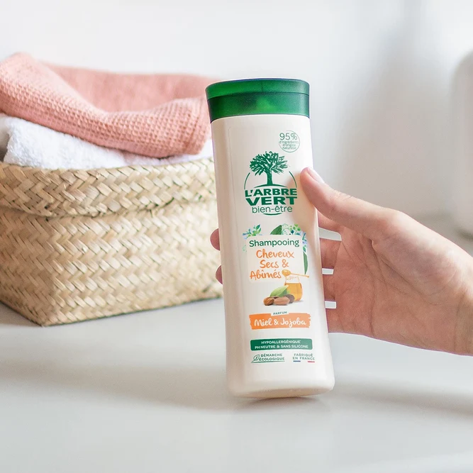 Une main tenant un flacon de 250 ml de shampooing L'Arbre Vert bien-être Cheveux Secs & Abîmés au parfum Miel & Jojoba, posé à côté d'une corbeille de serviettes.