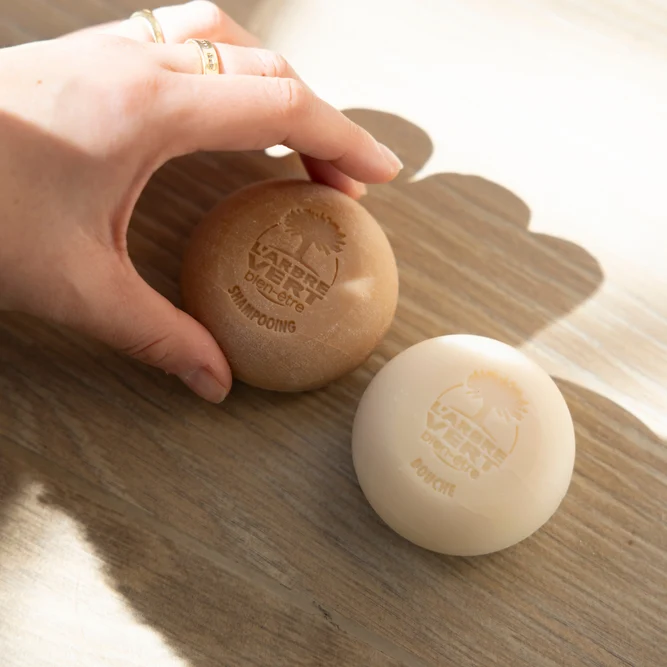 Une main attrapant un galet de shampooing solide L'Arbre Vert bien-être posé à côté d'un galet de produit douche solide sur une surface en bois.