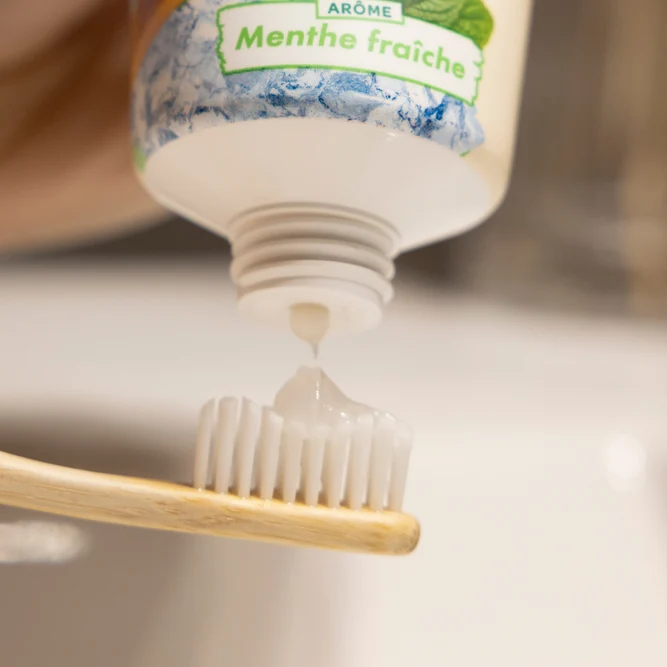 Texte alternatif (alt)  Gros plan sur l'application d'une noisette de dentifrice au gel blanc sur la tête d'une brosse à dents en bois, depuis un tube au parfum Menthe fraîche.