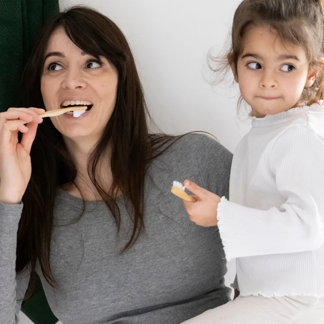 Une mère et sa jeune fille se brossant les dents ensemble avec des brosses à dents en bois dans la salle de bain.