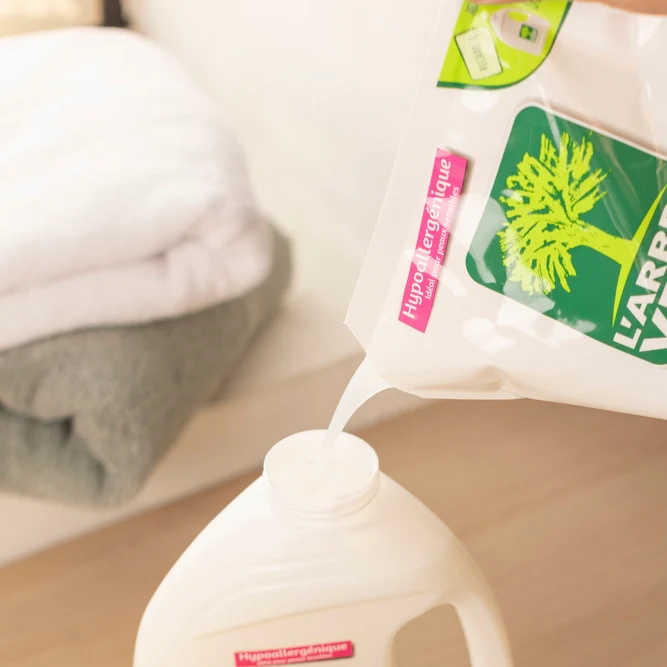 Texte alternatif (alt)  Remplissage d'un flacon blanc de lessive avec le contenu d'une sachet-recharge hypoallergénique L'Arbre Vert près d'une pile de serviettes.