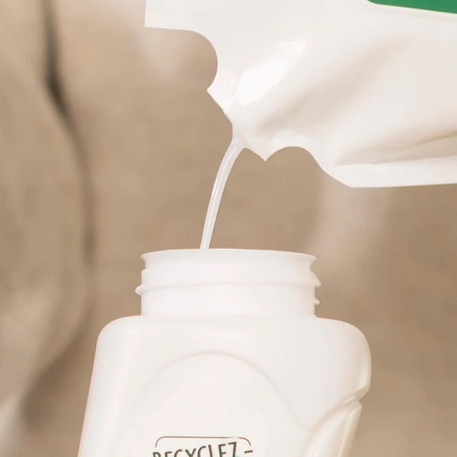 Gros plan sur le versement d'un liquide adoucissant blanc depuis un bec verseur de sachet-recharge dans le goulot d'un flacon blanc L'Arbre Vert.