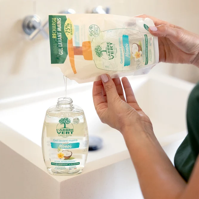 Une personne remplissant un flacon pompe transparent de Gel lavant mains L'Arbre Vert bien-être parfum Monoï à l'aide d'un sachet-recharge écologique (300 ml).