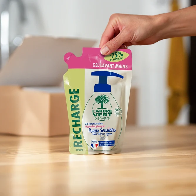 Een hand die een navulzak van 300 ml L'Arbre Vert handwasgel voor gevoelige huid op een houten tafel plaatst.